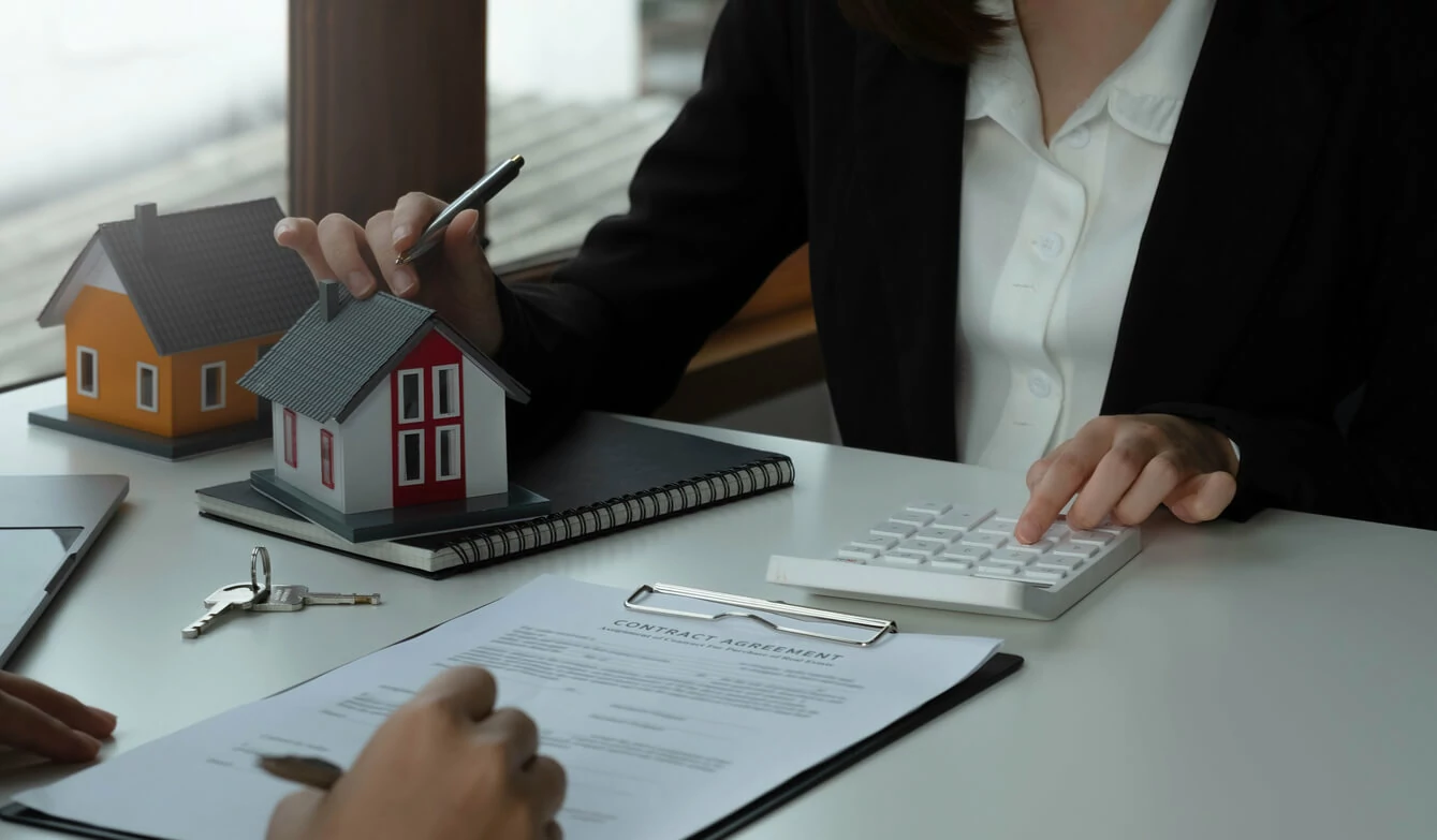 Femme avec une main posée sur une maquette de maison et l'autre sur une calculatrice devant un homme qui s'apprête à signer un document.