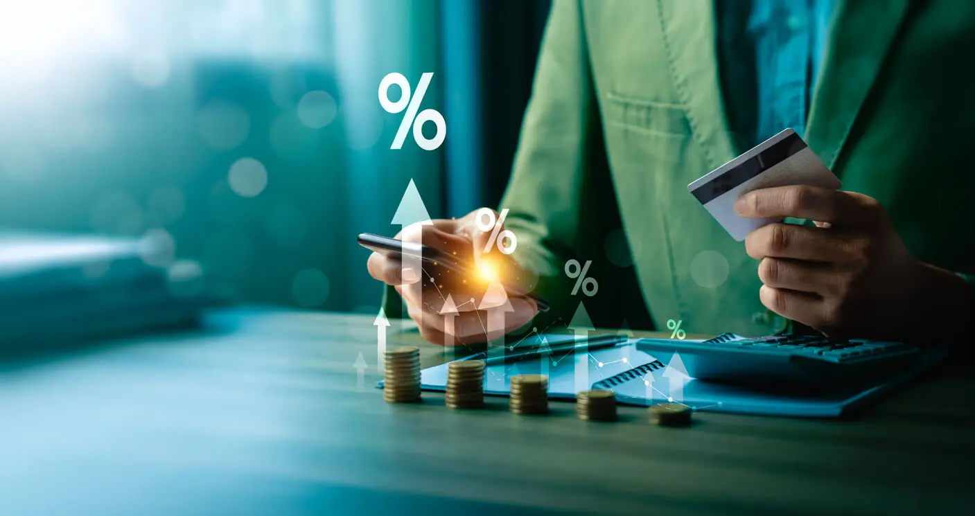 Homme tenant un smartphone et une carte de crédit devant plusieurs piles de pièces de monnaie, un bloc-notes et une calculatrice.