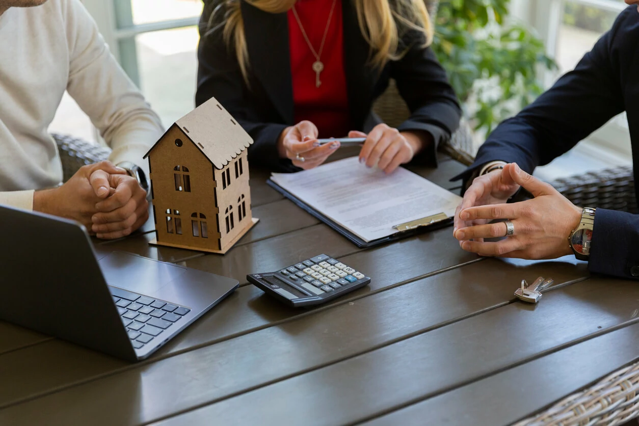 Trois personnes autour d'une table avec un ordinateur portable, une calculatrice, une maquette de maison, un document et une clé.