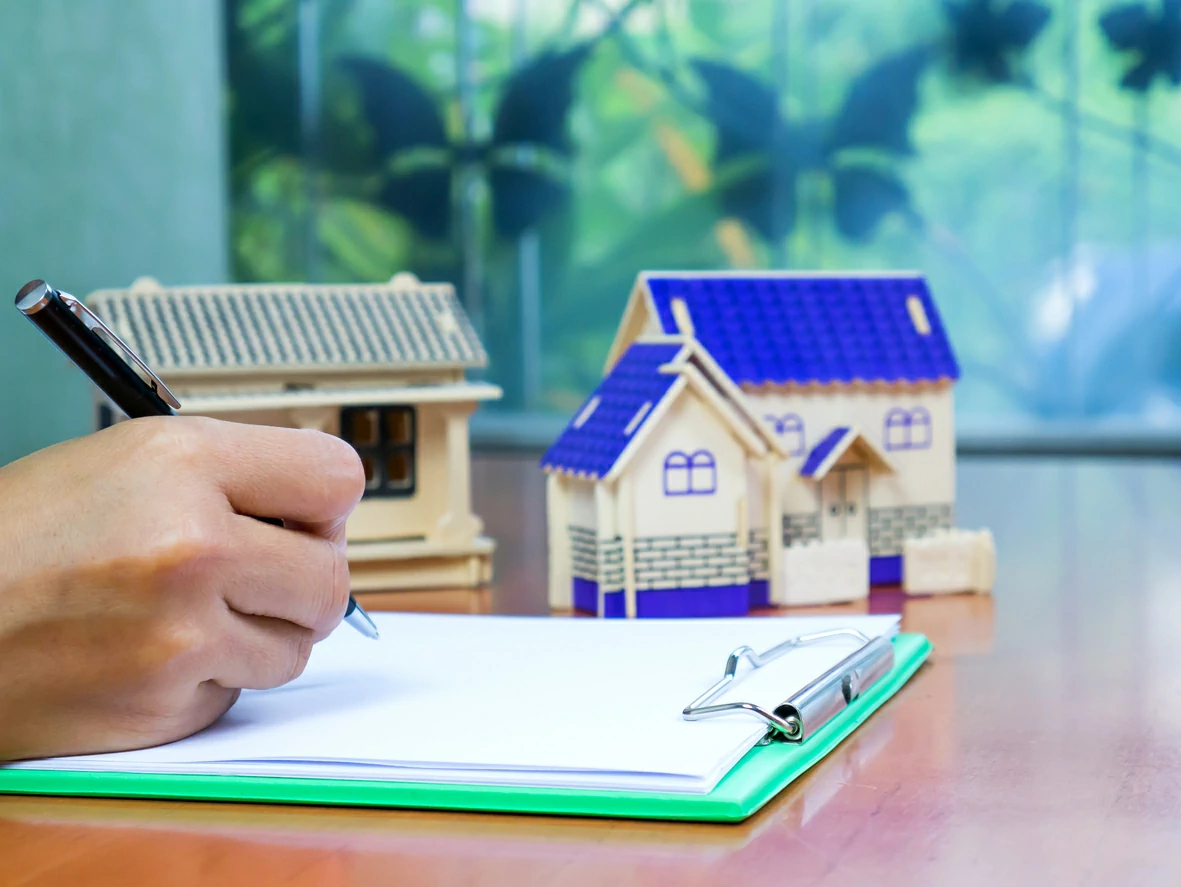 Main tenant un stylo qui écrit sur un document et trois petites maisons en arrière-plan