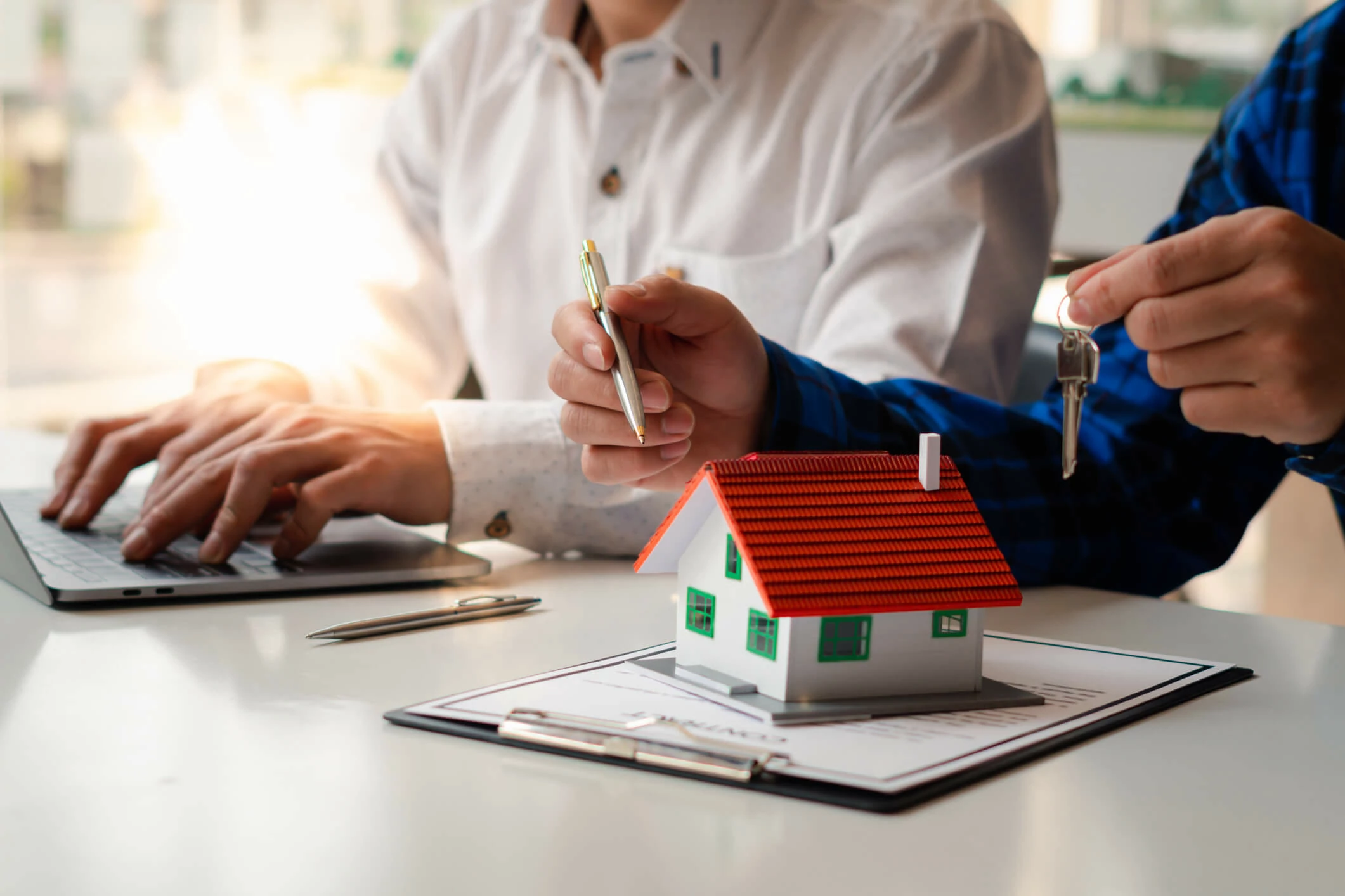 Homme tenant une clé et un stylo au-dessus d'une petite maison et d'un document à côté d'un homme devant un ordinateur