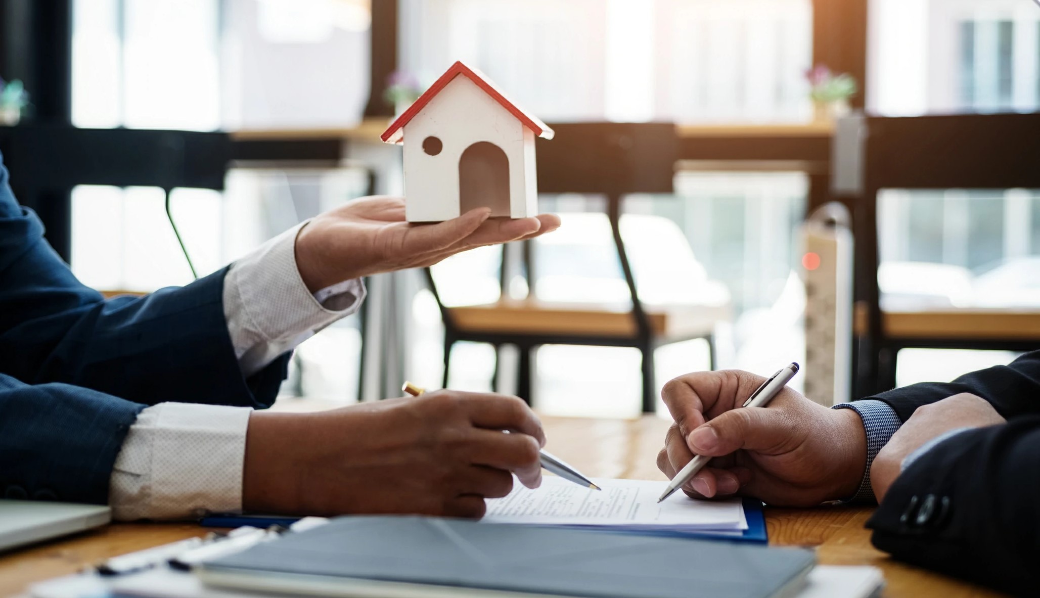 Homme tenant un petite maison en bois d'une main et un stylo de l'autre devant un autre homme qui s'apprête à signer un document.