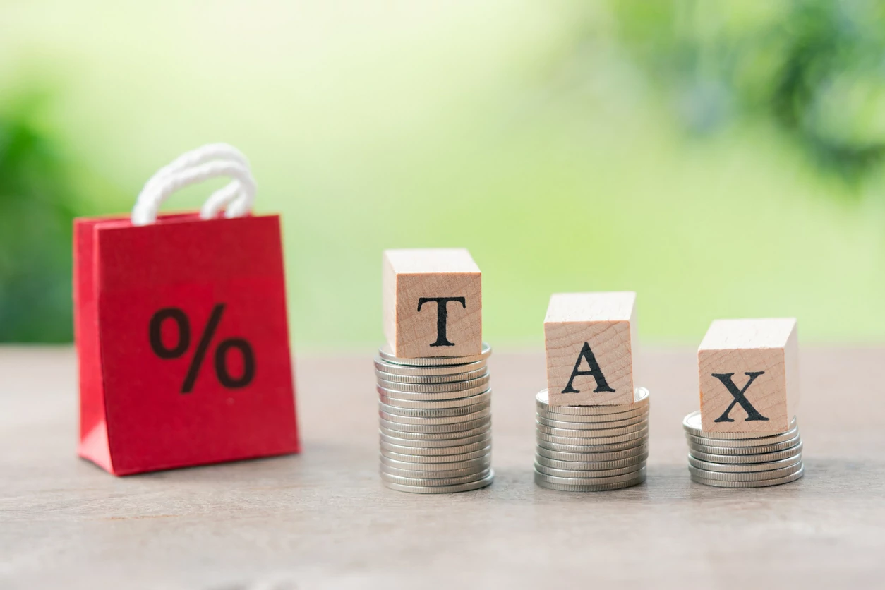 Trois blocs de bois avec le mot tax posés sur trois piles de pièces de monnaie et un petite sac en arrière-plan.