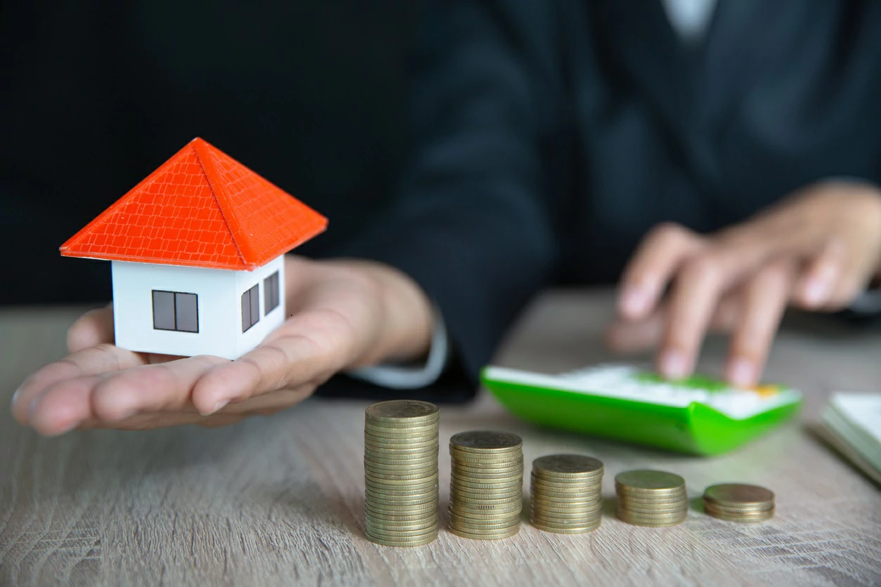 Personne tenant une petite maison dans la main gauche et utilisant une calculatrice dans la main droite devant des piles de pièces de monnaie.