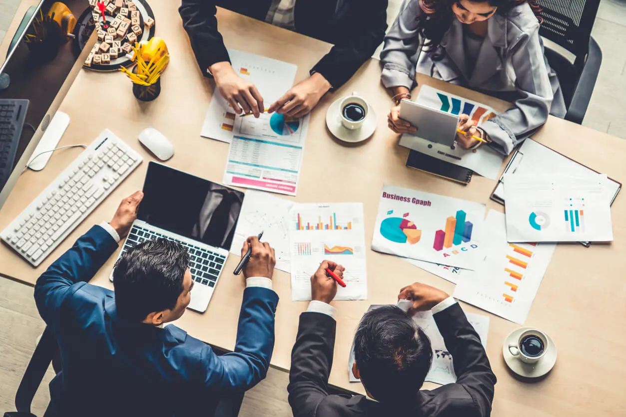 Quatre personnes autour d'une table remplie de documents, des ordinateurs des tasses de cafés et d'autres fournitures.
