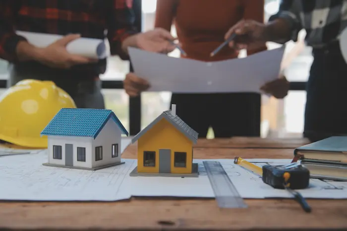 Deux maquettes de maison posées au-dessus de plusieurs documents et trois personnes consultant des plans en arrière-plan.