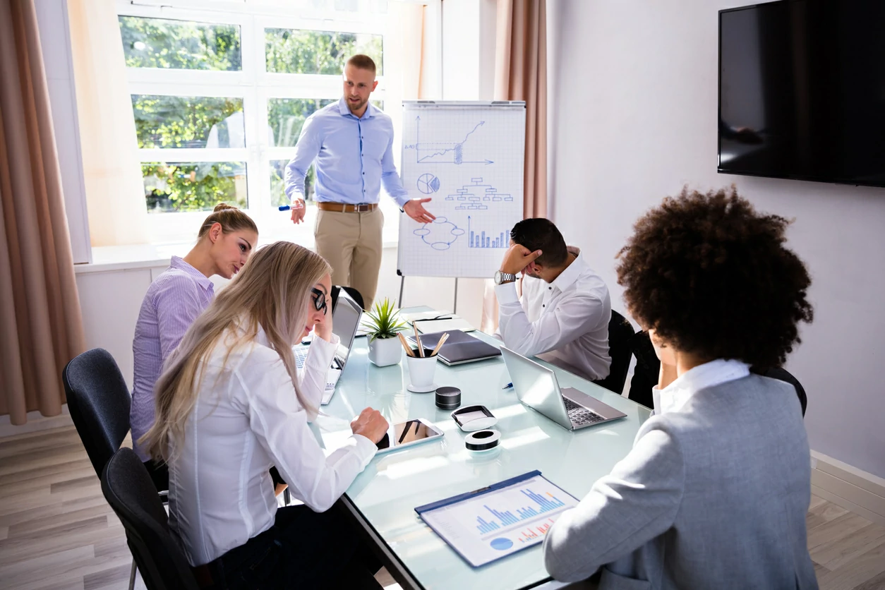 Homme devant un tableau blanc et quatre personnes assises autour d'une table avec des ordinateurs portables et des documents