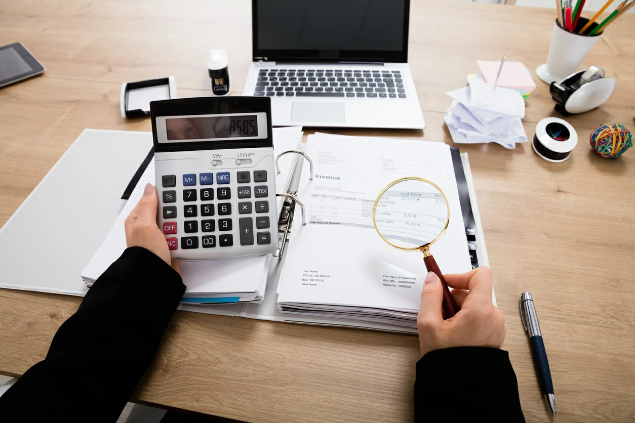 Main tenant une calculatrice au-dessus de plusieurs documents, d'ordinateur portable et d'autres fournitures de bureau.