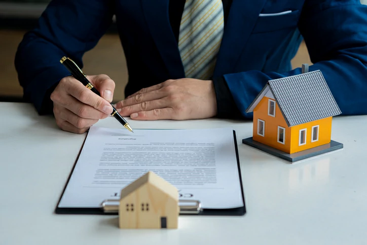 Homme en costard signant un document avec un stylo à côté de deux petites maisons.