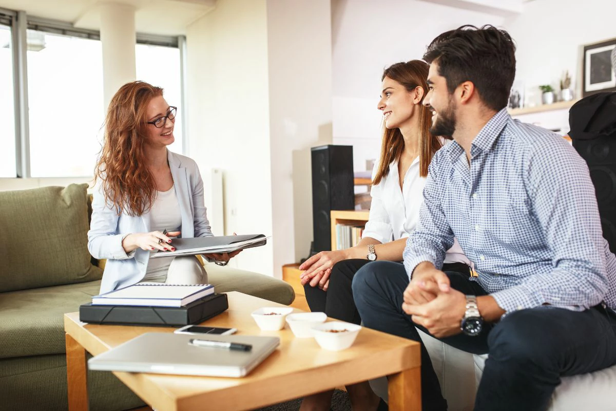 Femme tenant un document devant un couple souriant autour d'une table remplie de document et de nourriture.
