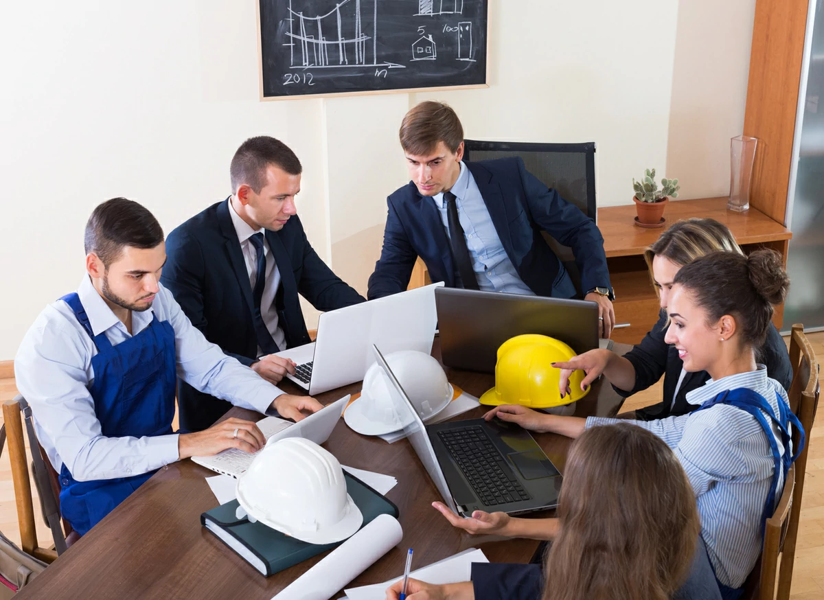 Six personnes en réunion autour d'une table remplie de documents, d'ordinateurs portables et des casques de chantier. 