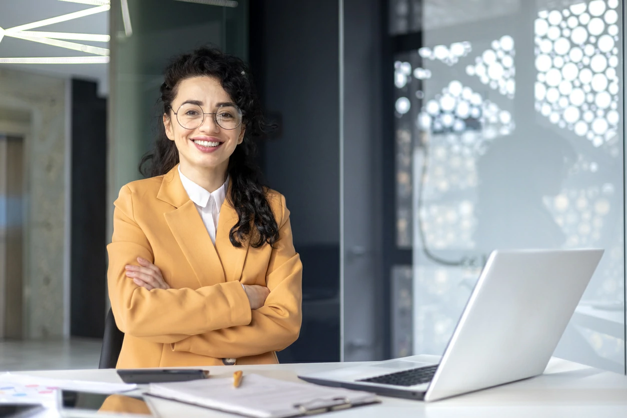 Une professionnelle à lunettes devant un bureau avec des documents et un ordinateur portable.