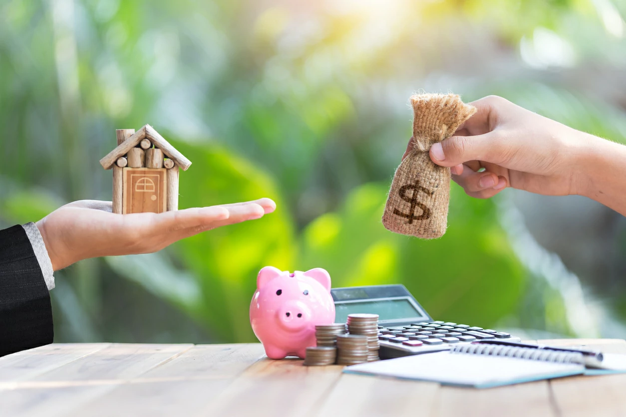 Personne tenant une petite maison en bois et une autre personne en face tenant une petite bourse d'argent.