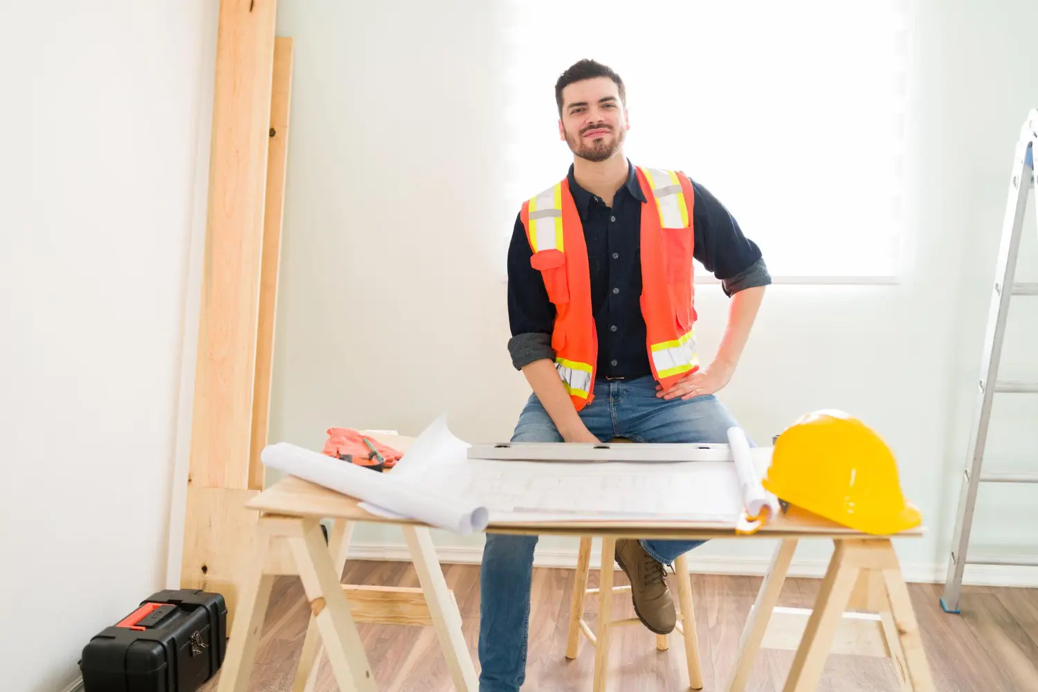 Professionnel du BTP avec un gilet orange devant une table remplie de documents divers et un casque de chantier.