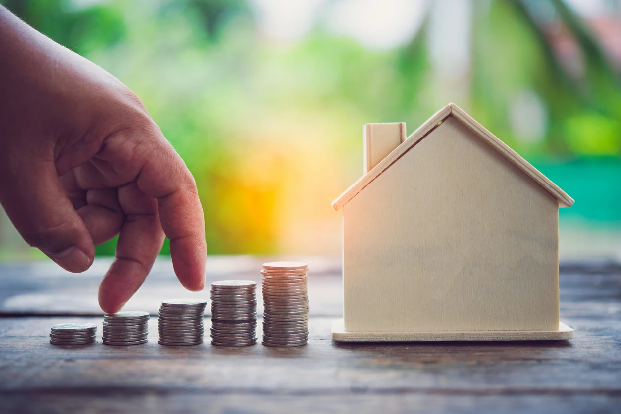 Main qui monte sur plusieurs piles de pièces de monnaie à côté d'une petite maison.