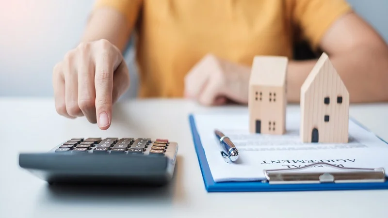 Main de femme utilisant une calculatrice à côté de documents et de maison en bois. 
