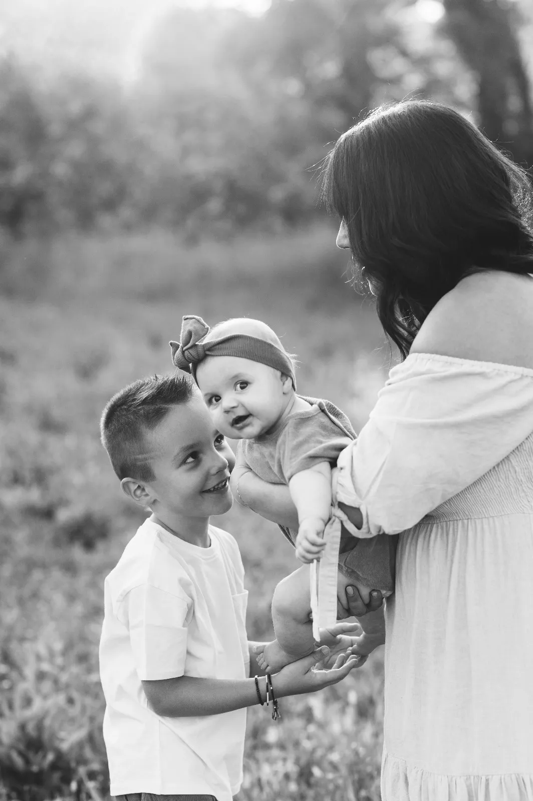 Sessione bebè in esterna in bianco e nero: il fratello maggiore guarda la sorellina in braccio alla mamma