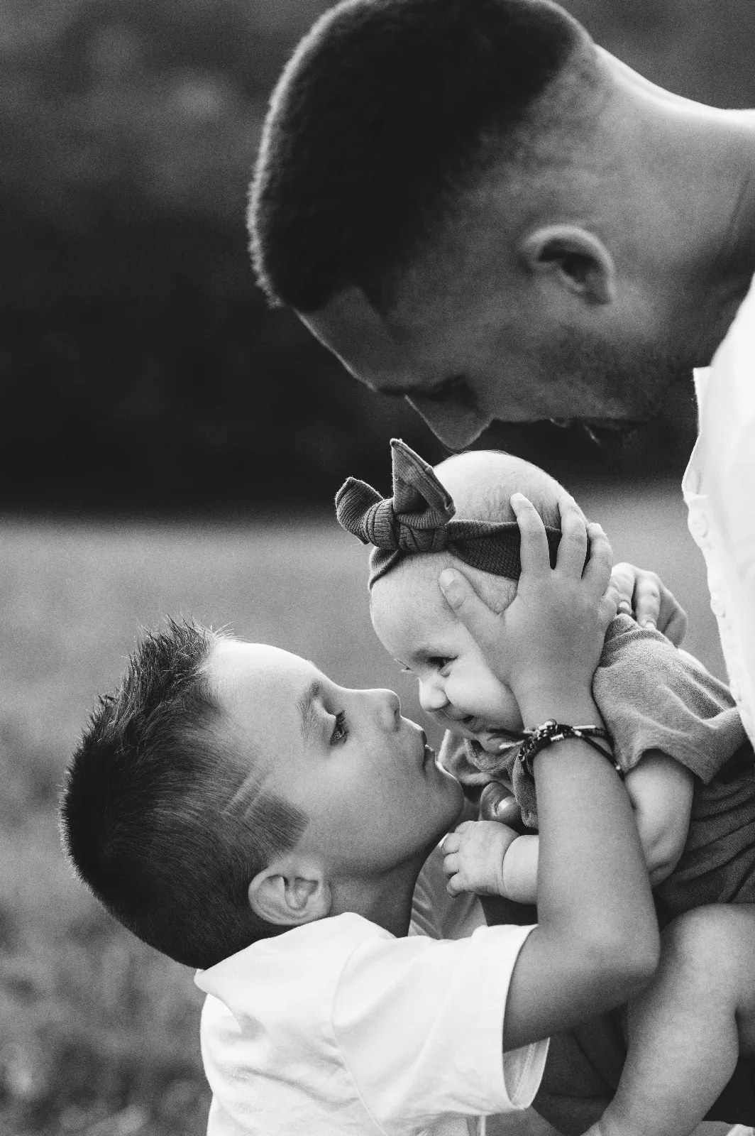 Sessione bebè in esterna in bianco e nero: papà tiene in braccio la bimba mentre il fratello le dà un bacio