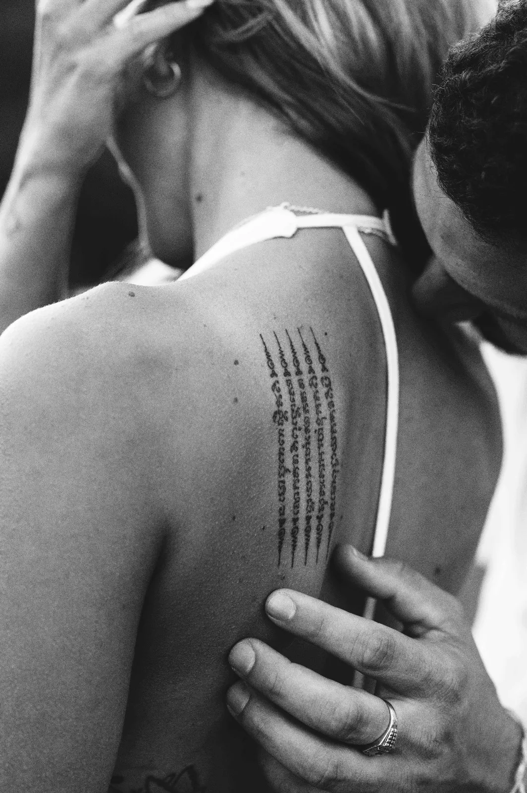 Dettaglio di coppia in bianco e nero: l'abbraccio da dietro e il tatuaggio sulla spalla di lei
