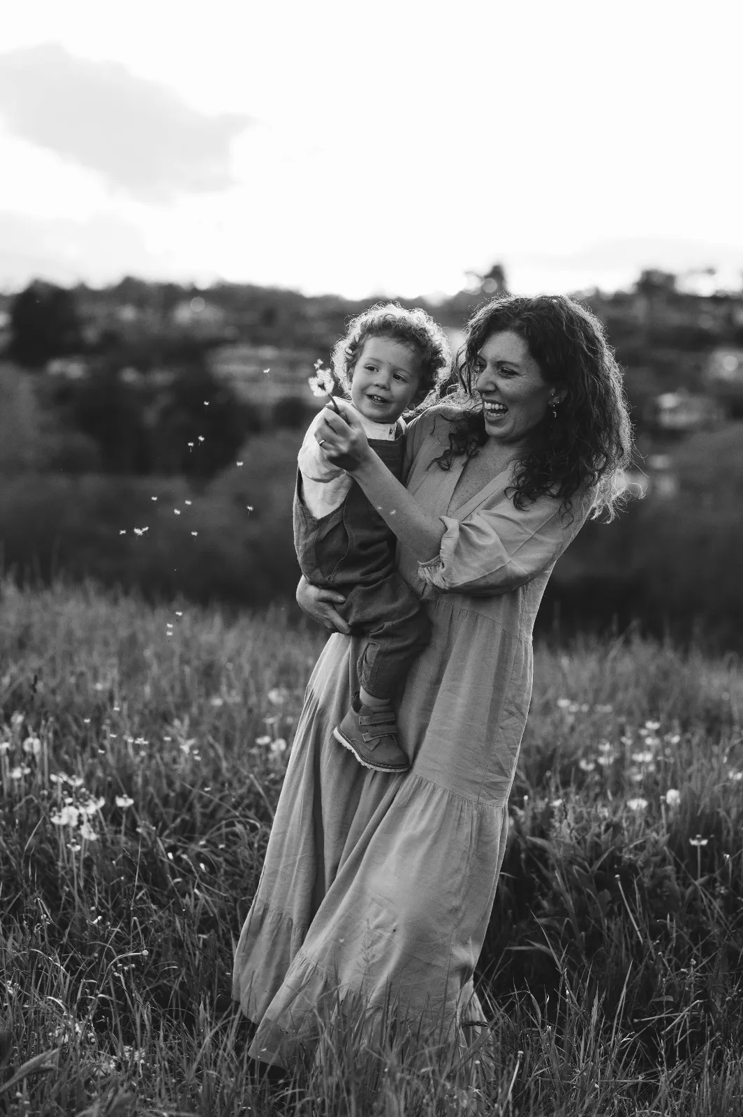 Servizio di famiglia in bianco e nero: mamma con il figlio in braccio che soffia un soffione in collina