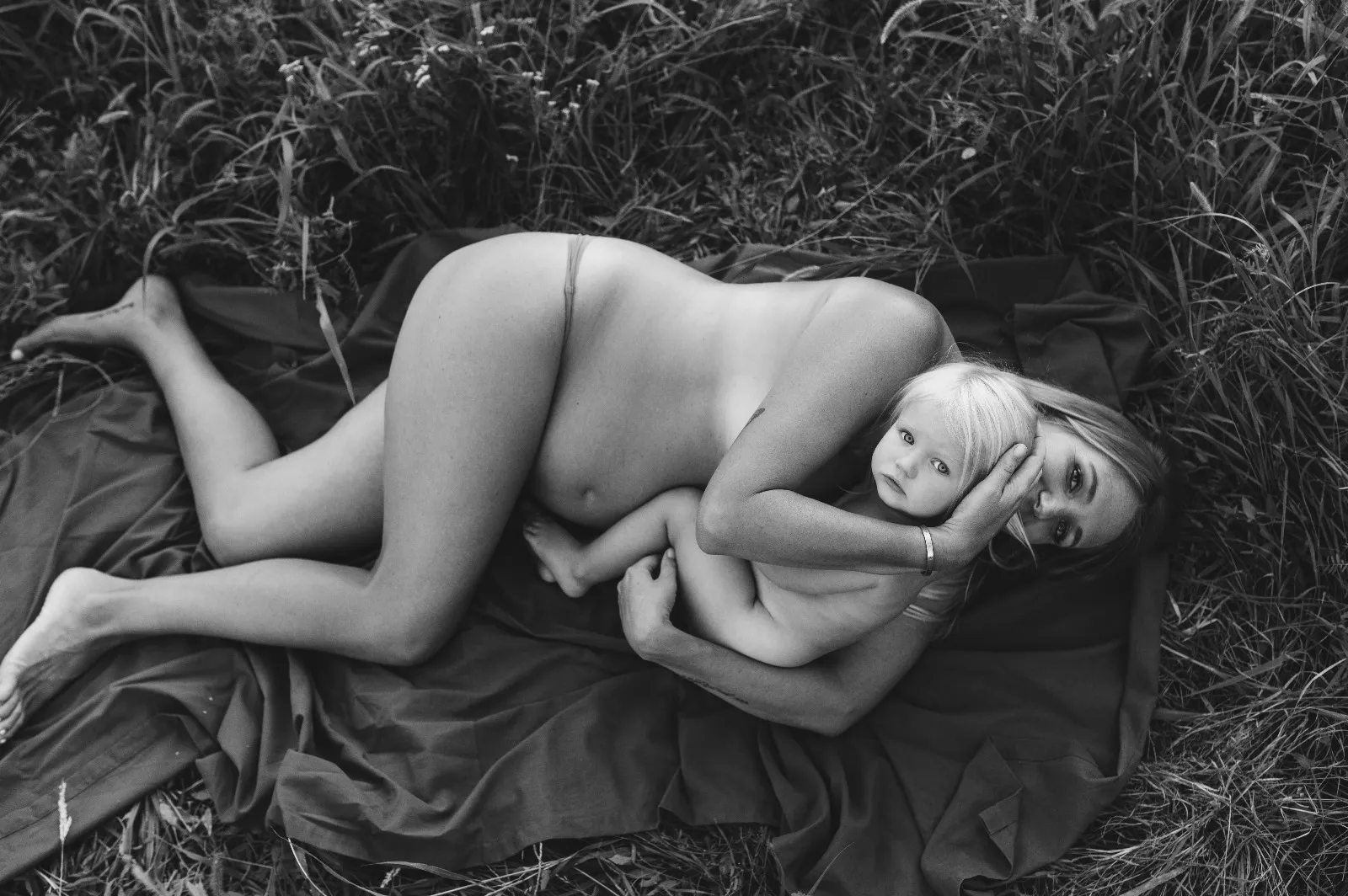 Servizio fotografico maternity in esterna: mamma in attesa distesa su un telo nell'erba abbraccia la figlia, in bianco e nero