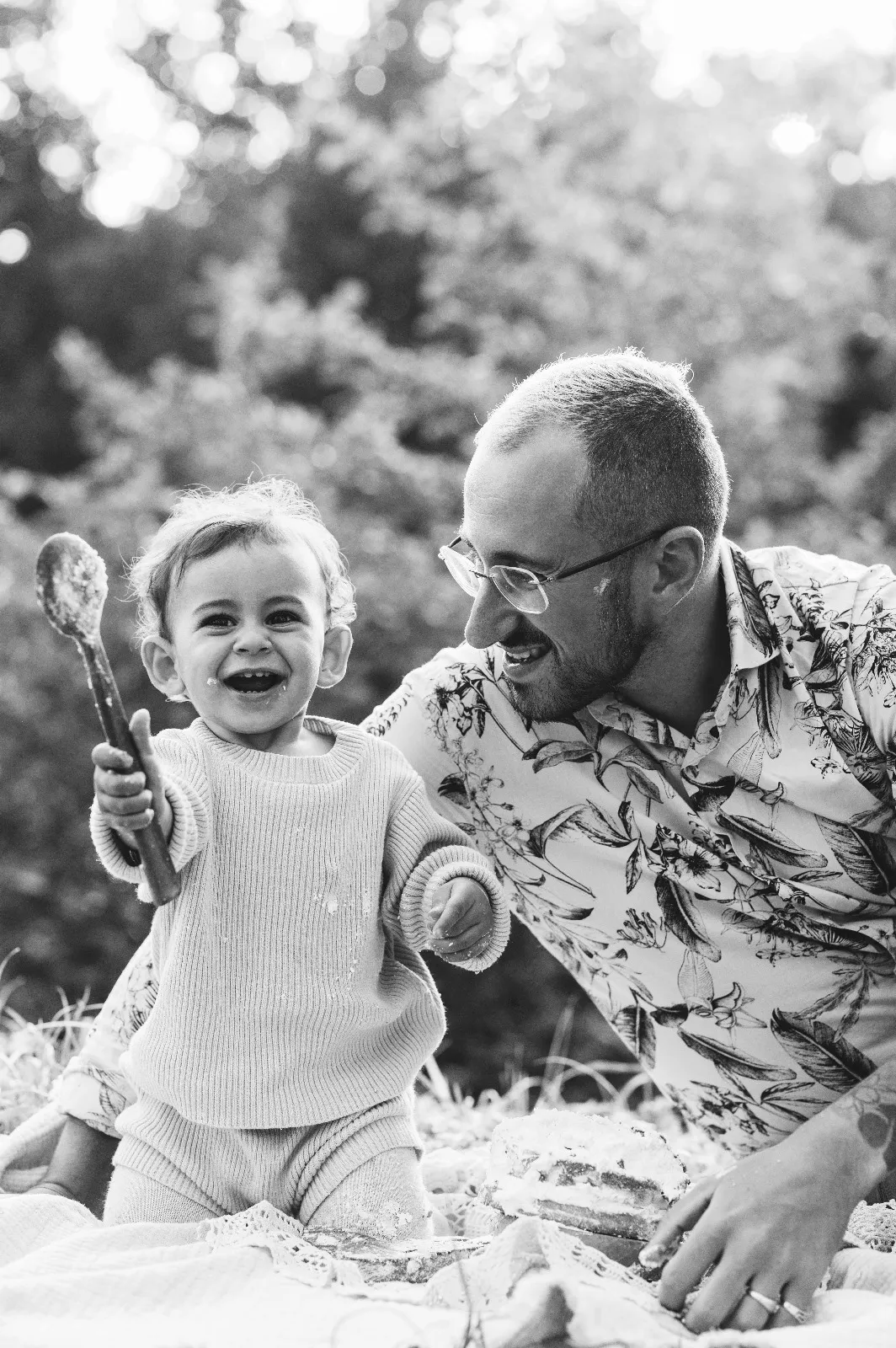 Sessione smash cake in esterna in bianco e nero: la bimba ride con il cucchiaio in mano accanto al papà