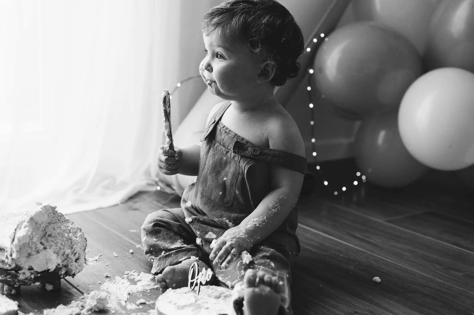 Servizio fotografico smash cake in studio: bimbo seduto con il cucchiaio di legno accanto alla torta distrutta, tra i palloncini, in bianco e nero