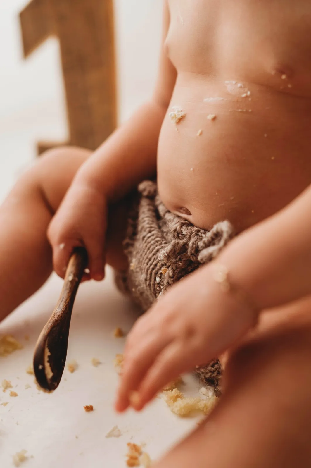 Dettaglio della sessione smash cake: le mani del bimbo con il cucchiaio tra le briciole di torta, accanto al numero uno in legno