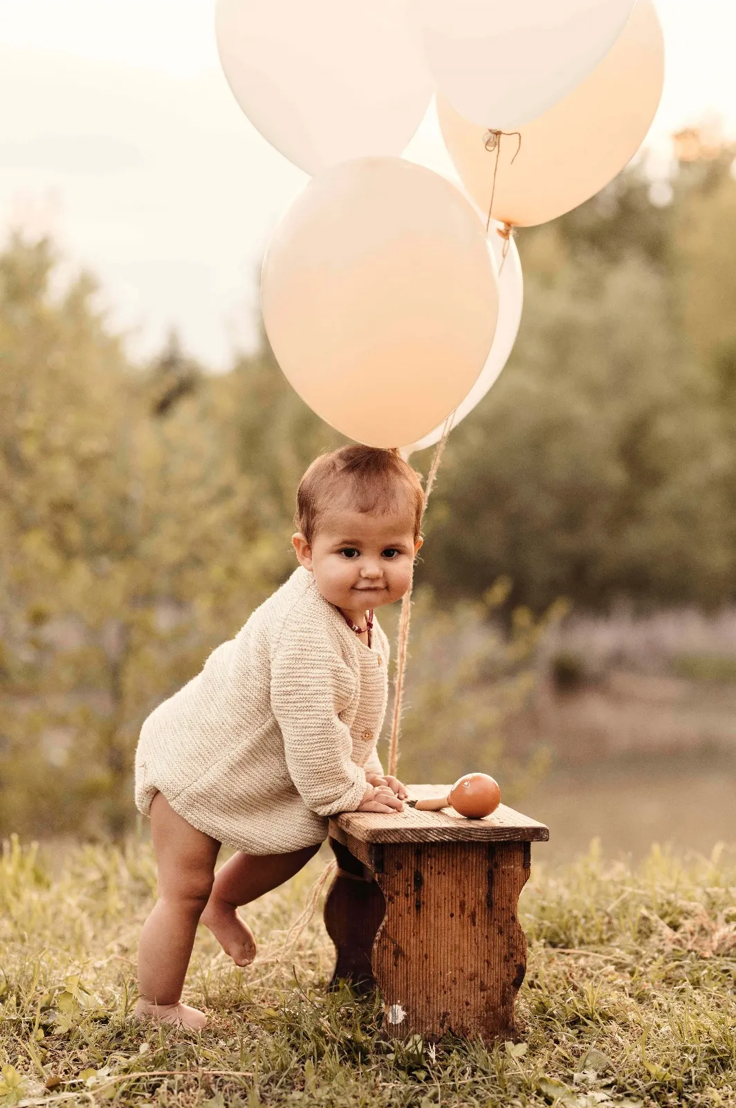 Servizio fotografico primo compleanno in esterna: bimba in piedi accanto allo sgabello di legno con i palloncini