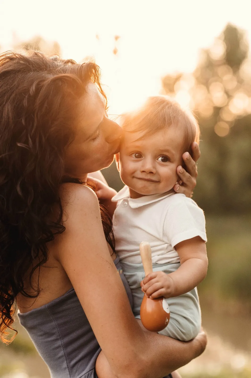 Servizio fotografico bebè al tramonto: mamma bacia la testa del bimbo che tiene un sonaglio