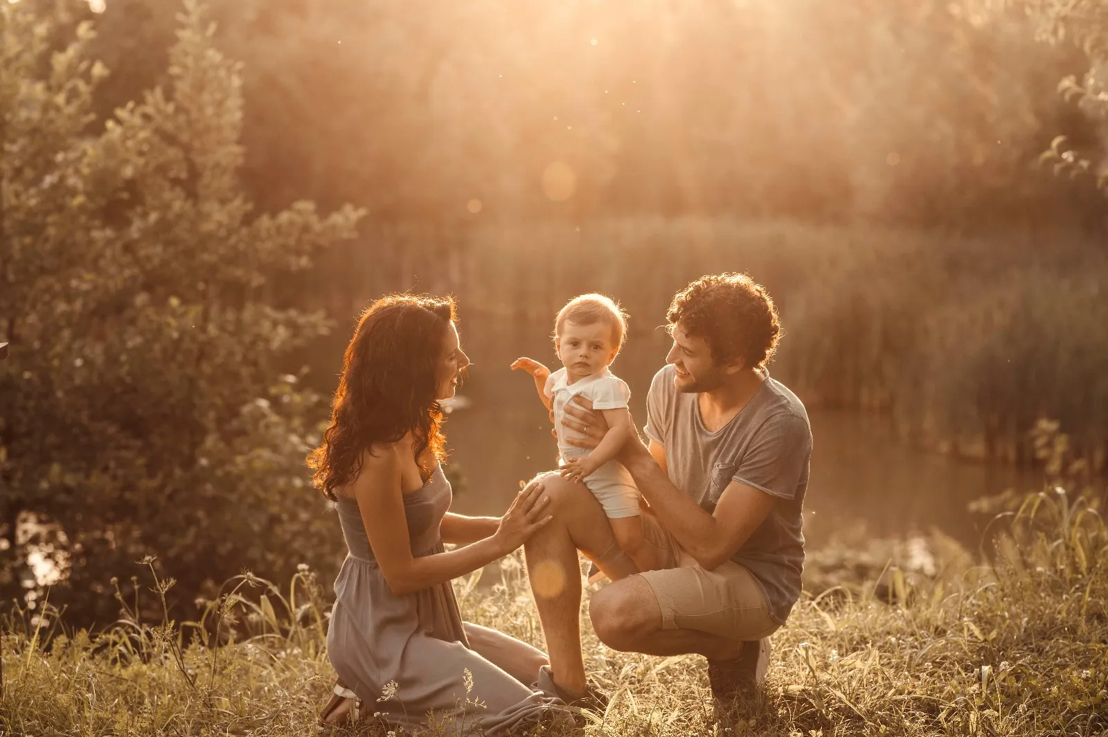 Servizio fotografico bebè in esterna al tramonto: mamma e papà accovacciati accanto al bimbo in piedi in un prato