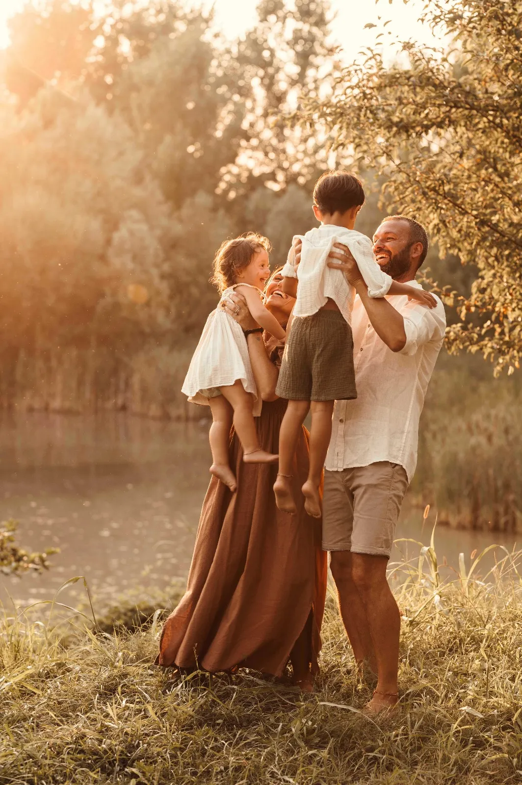 Servizio fotografico di famiglia al tramonto: genitori che sollevano i due bambini in braccio davanti a un laghetto