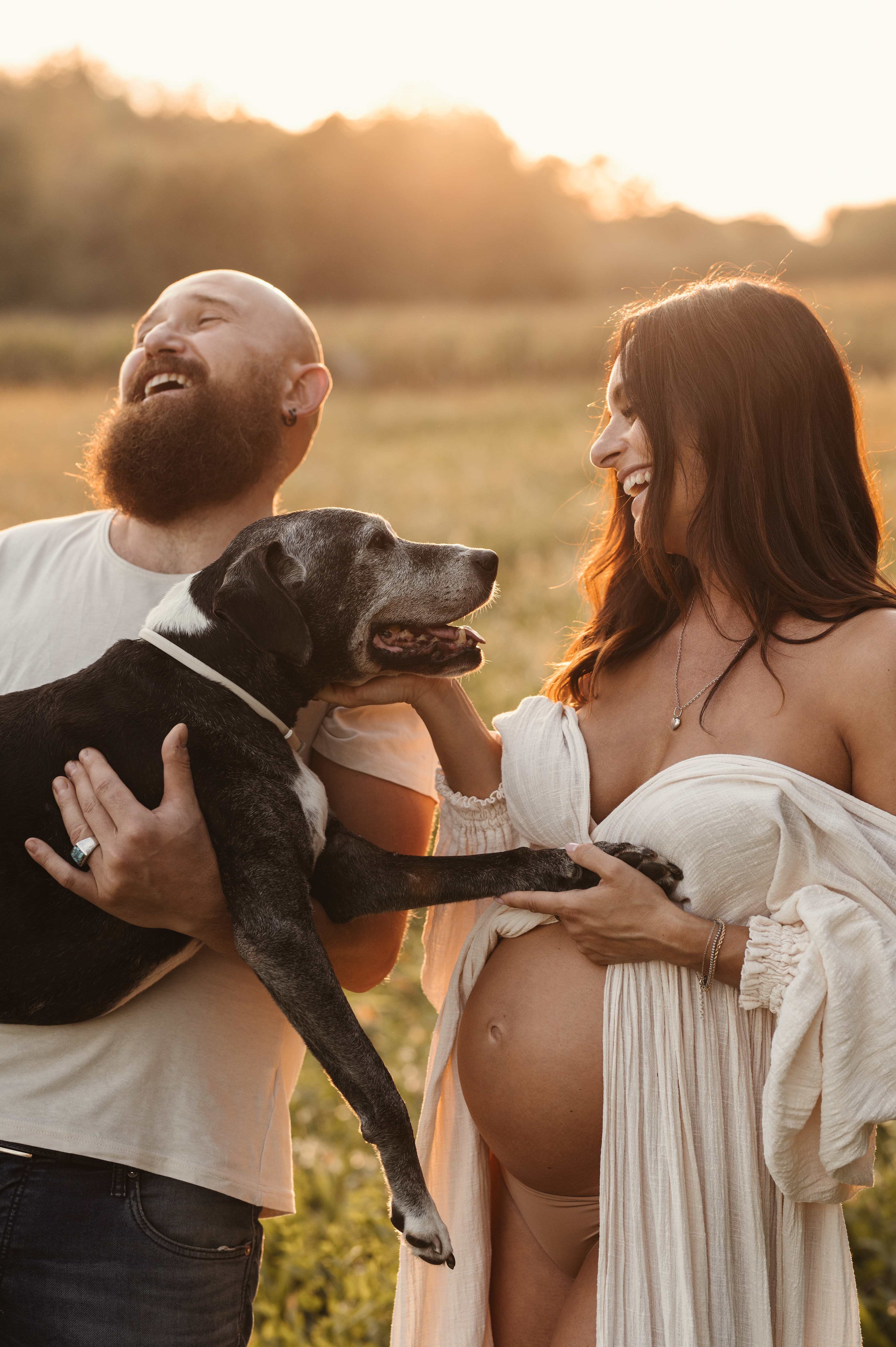 Sessione maternity al tramonto in un campo: coppia che ride insieme al cane, con il pancione in primo piano