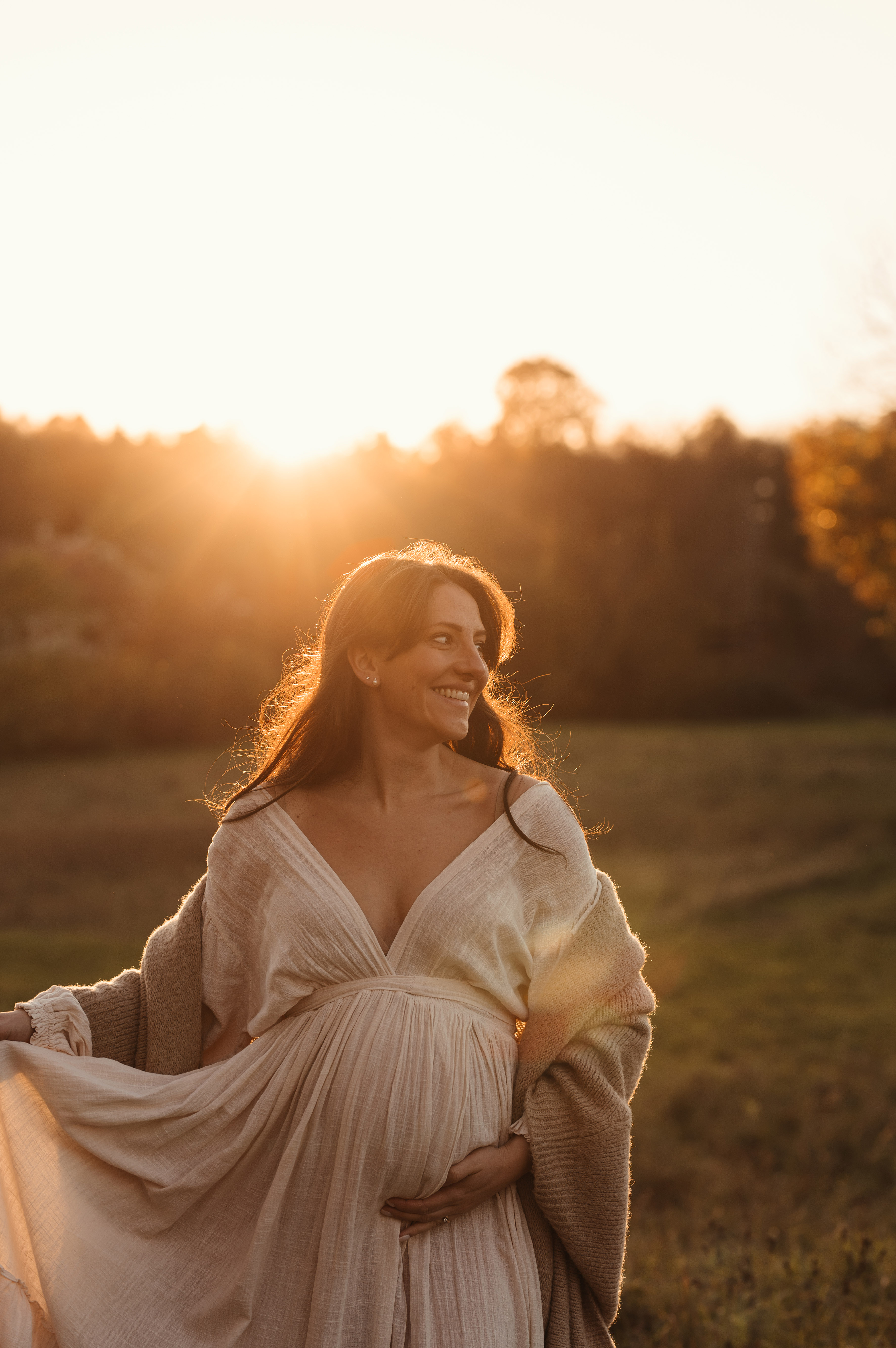 Sessione fotografica maternity al tramonto: futura mamma in abito lungo sorride controluce in un prato