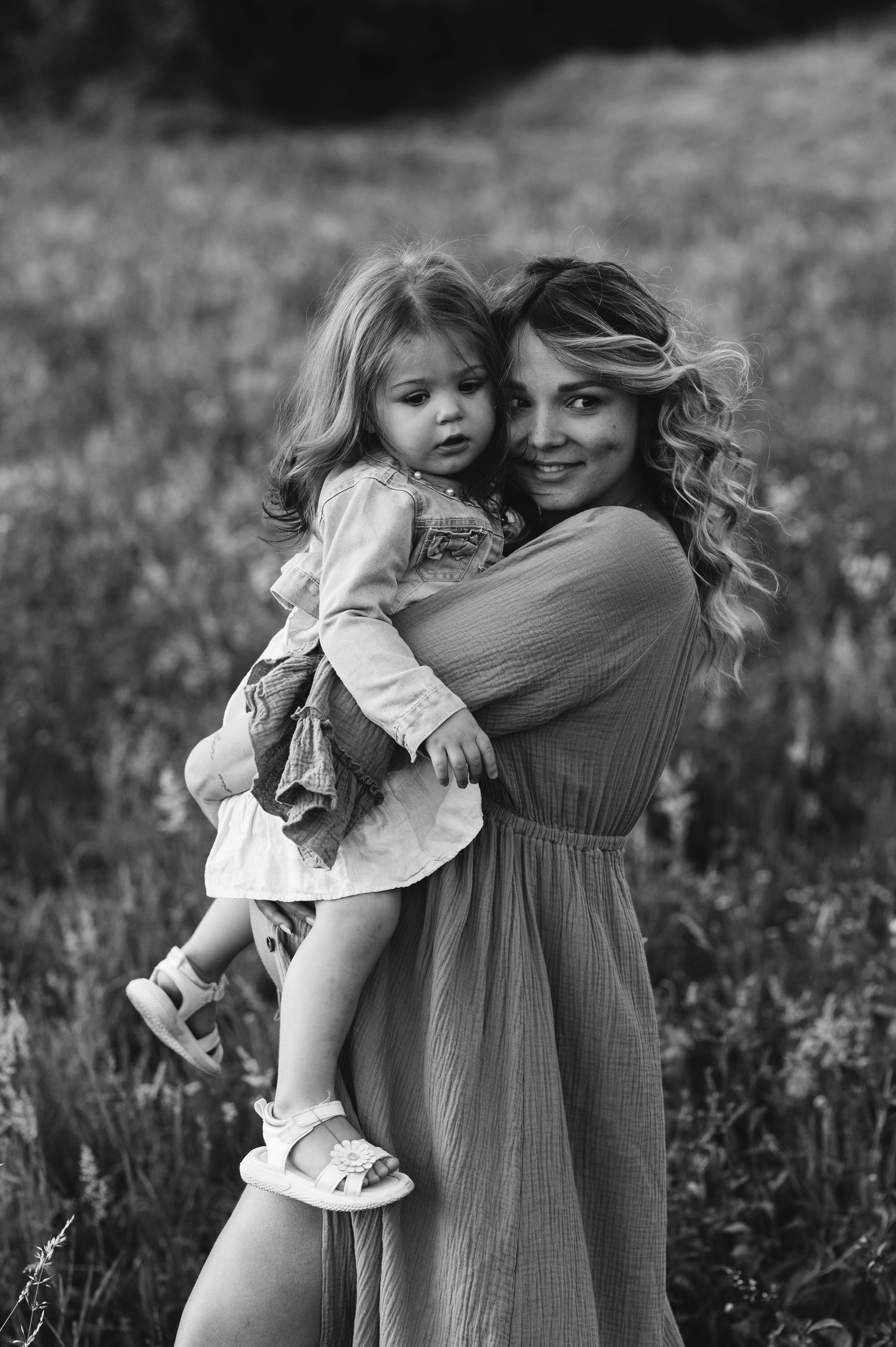 Servizio maternity in esterna: mamma in attesa tiene in braccio la figlia piccola in un prato fiorito, in bianco e nero