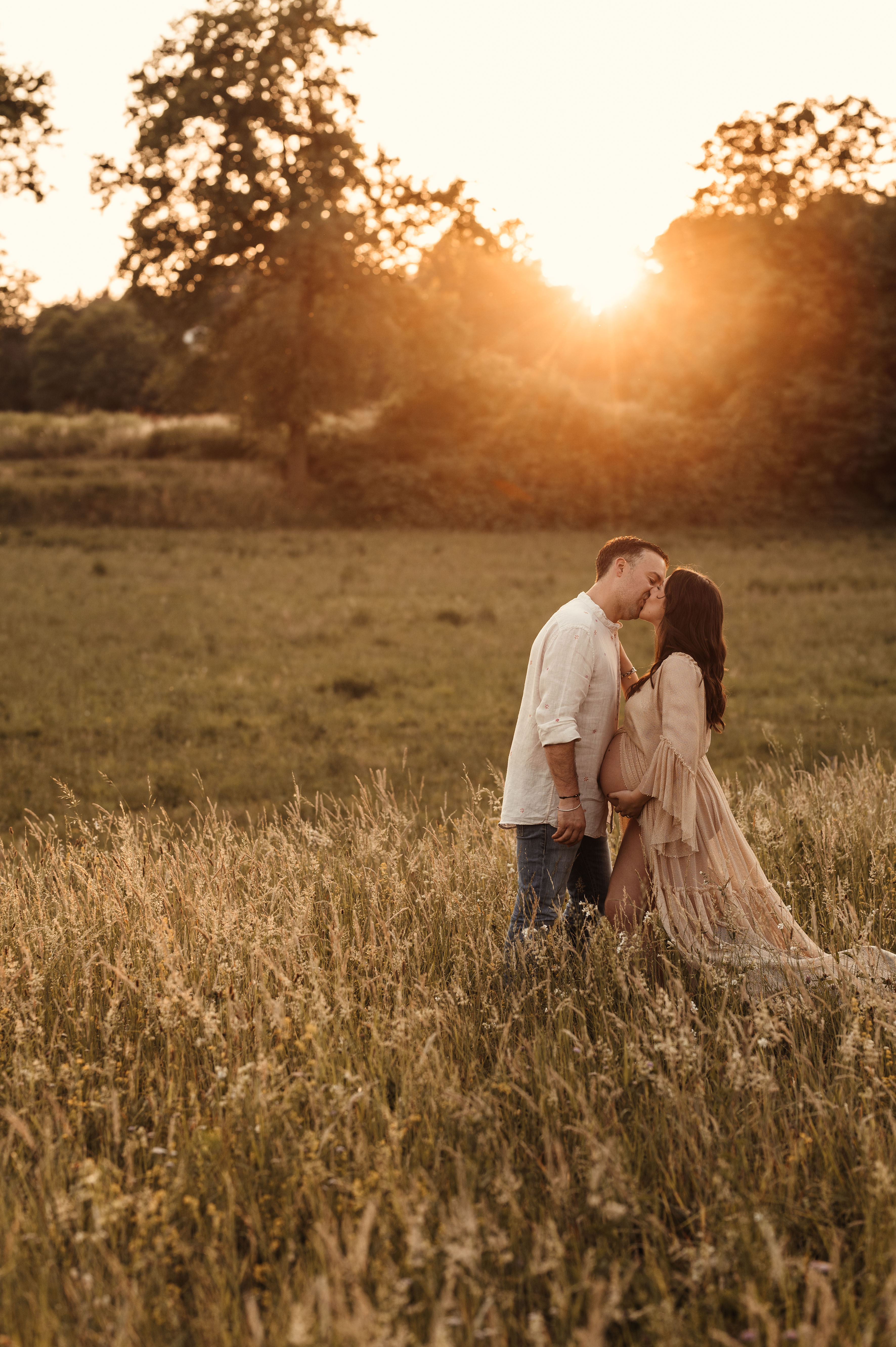 Foto di gravidanza al tramonto: coppia che si bacia in un campo di erba alta, luce dorata