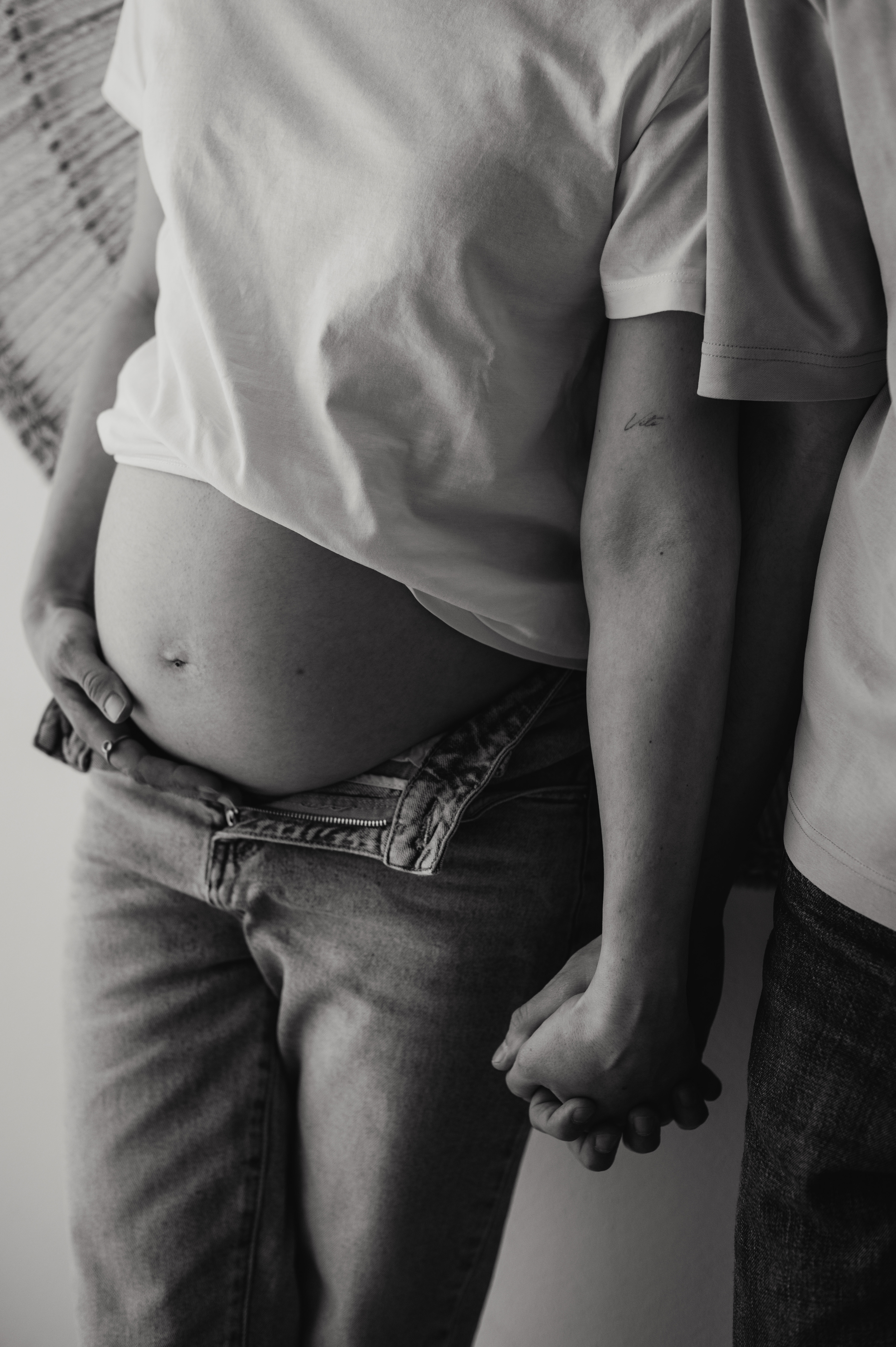 Foto maternity in studio: pancione in maglietta bianca e jeans, mano nella mano con il compagno, in bianco e nero
