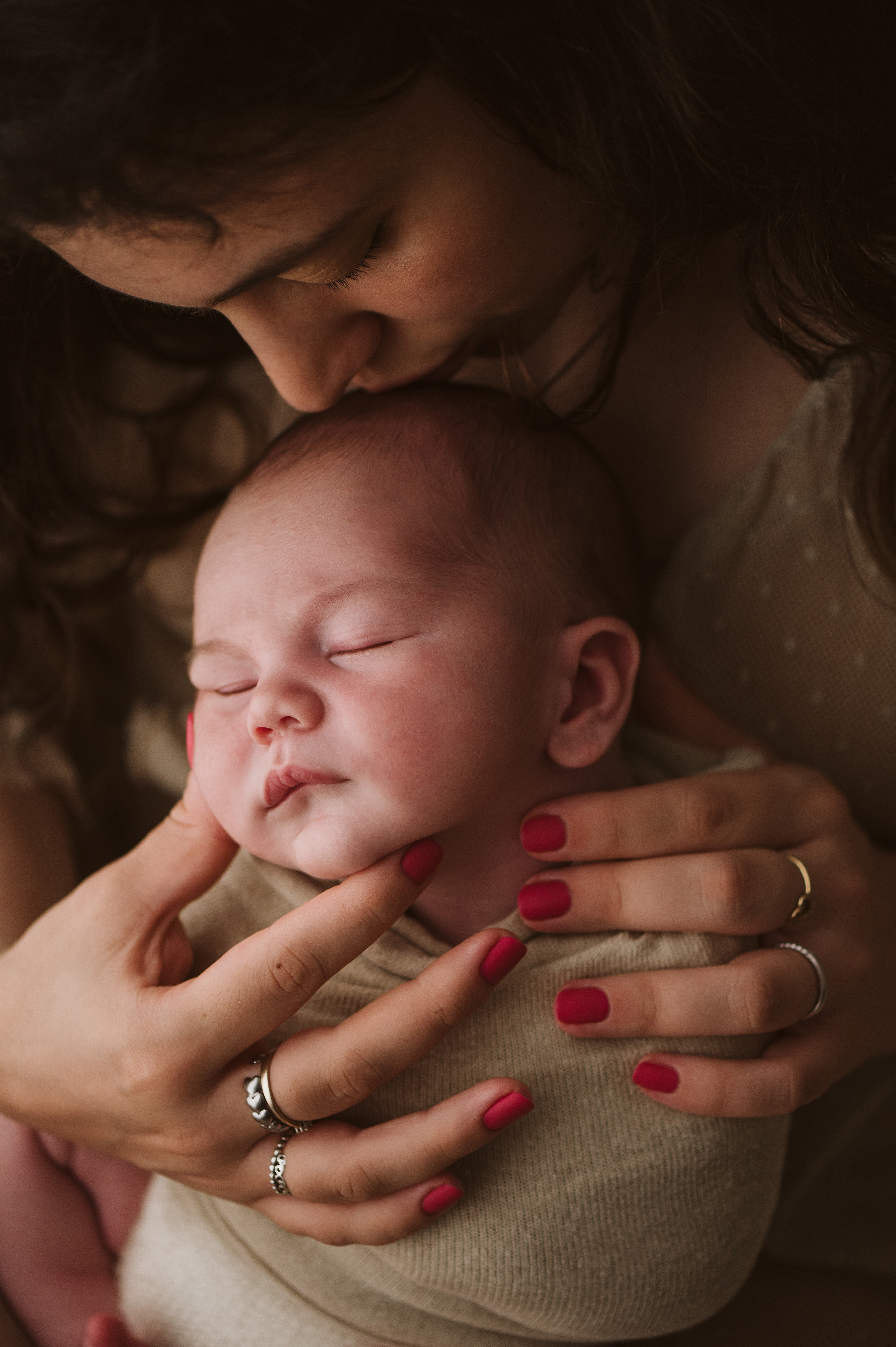 Sessione fotografica newborn: mamma bacia la fronte del neonato avvolto in una fascia, tonalità calde