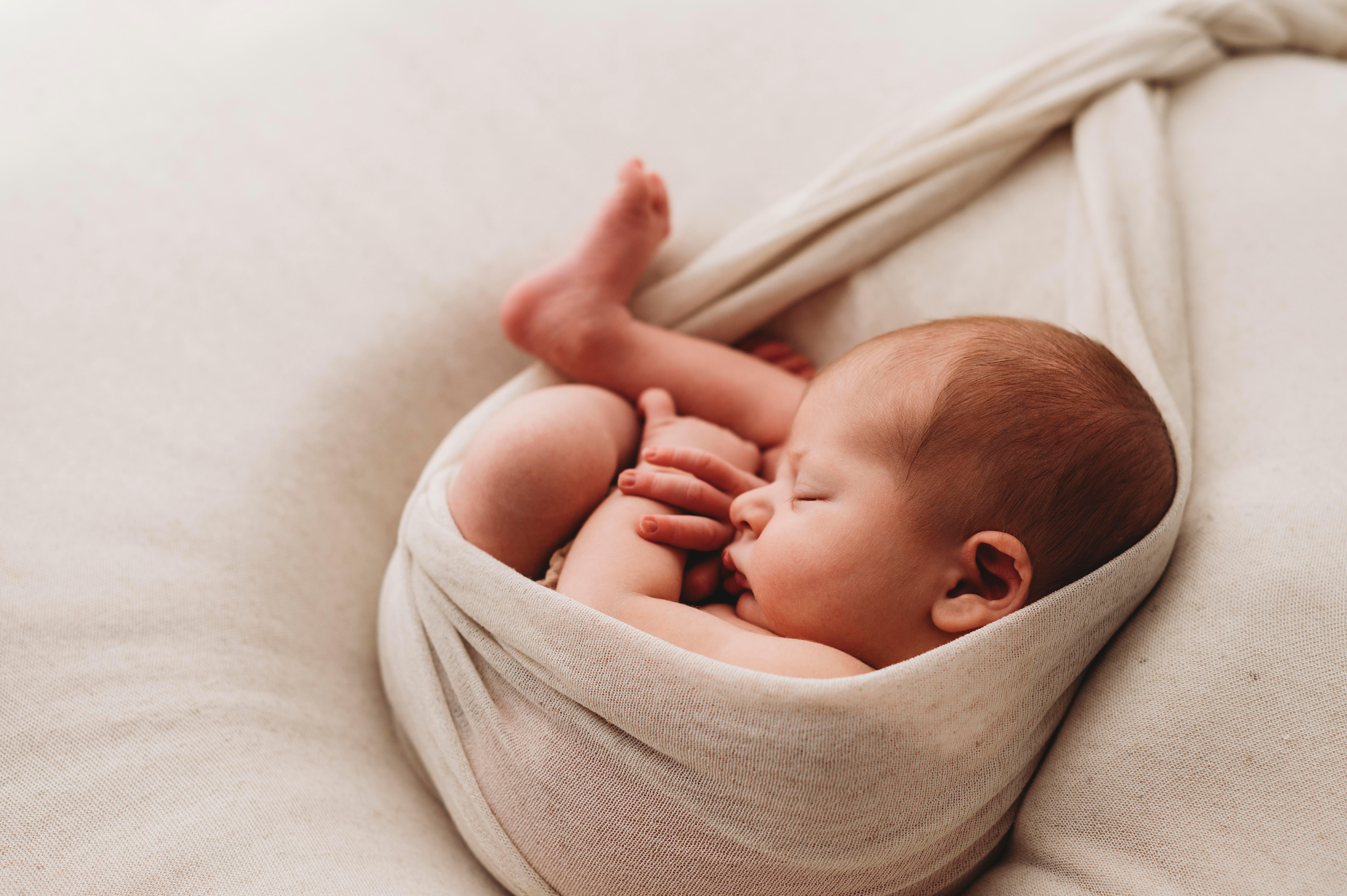 Neonato avvolto in una fascia beige mentre dorme su una coperta morbida, sessione newborn in studio
