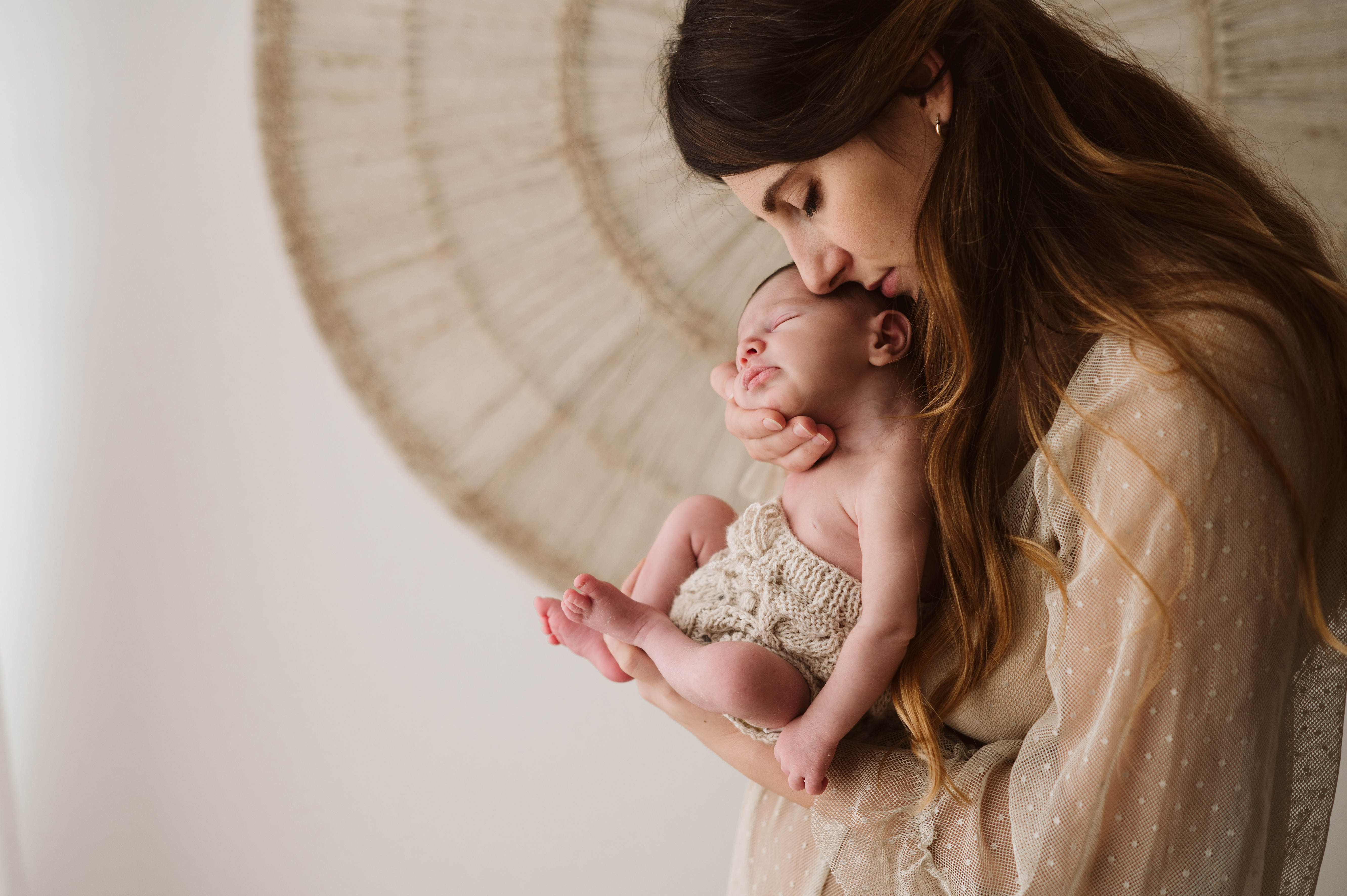 Servizio fotografico newborn in studio: mamma in piedi bacia la testa del neonato che tiene in braccio, luce naturale