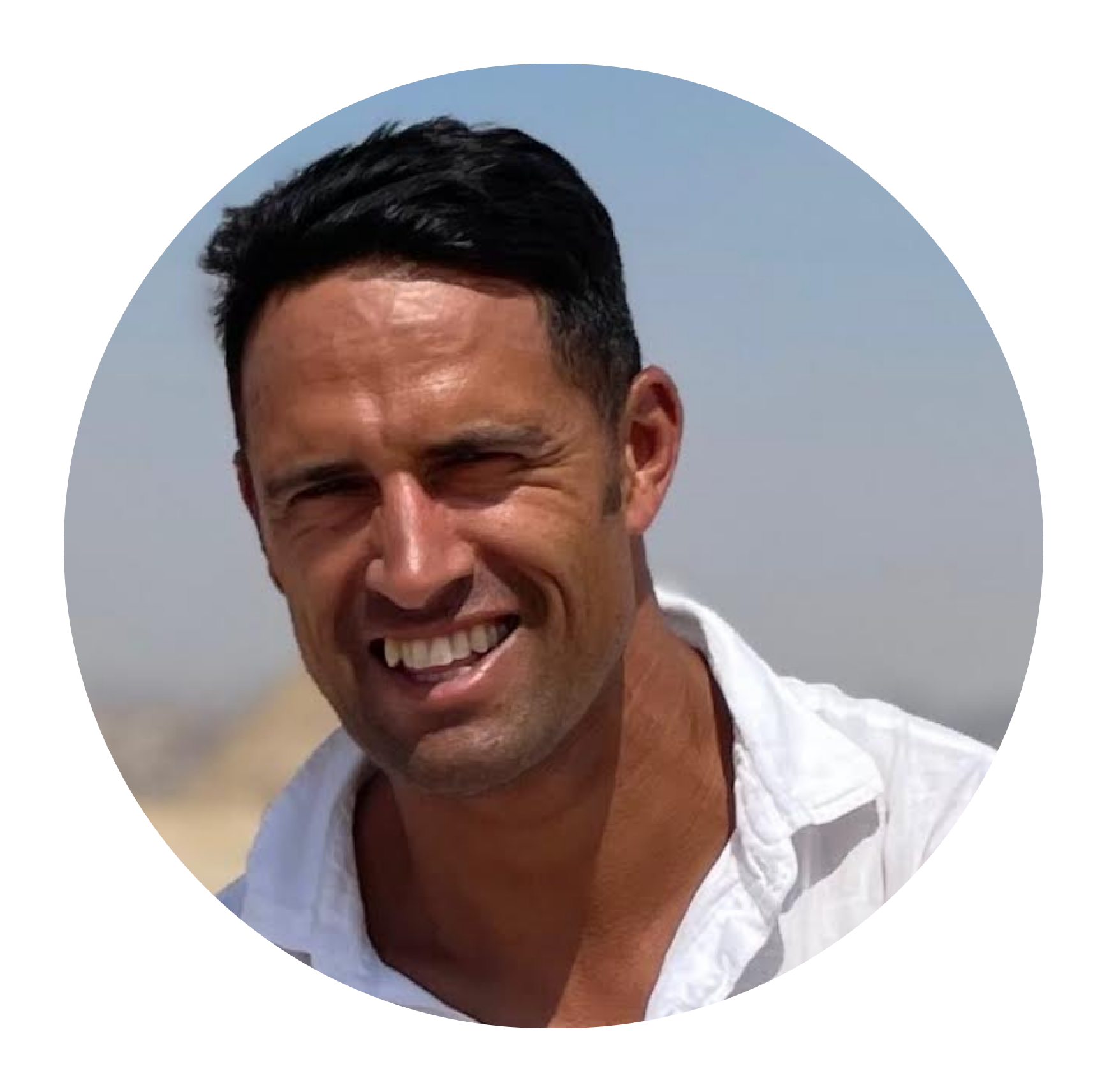 Smiling man with short dark hair wearing a white shirt against a clear sky background.