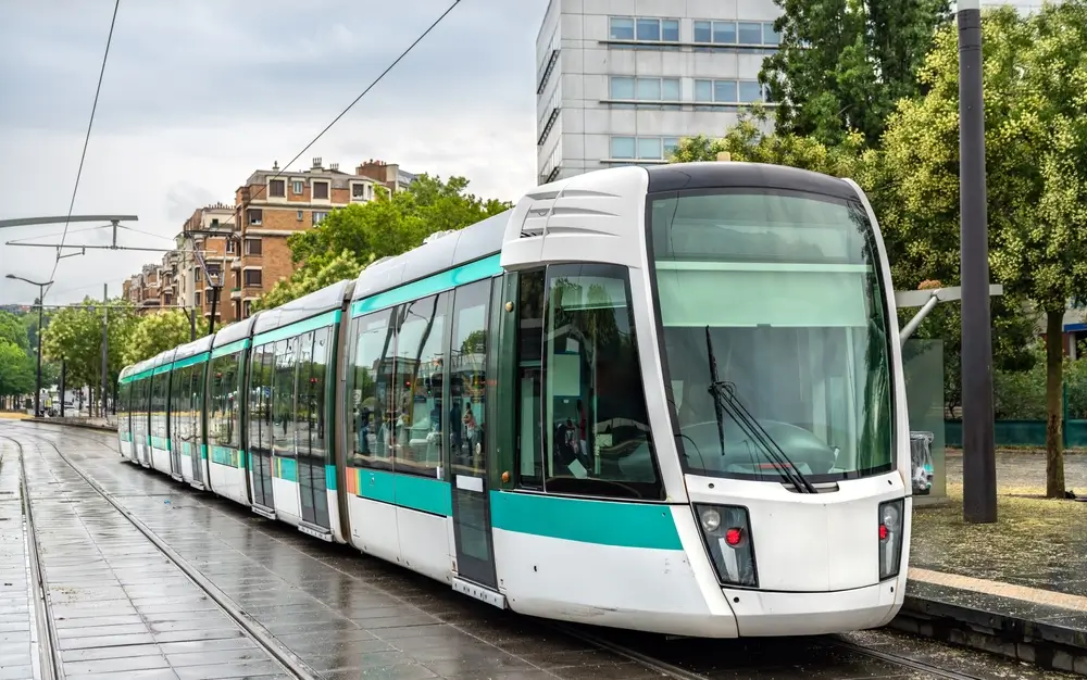 Un train à grande vitesse (TGV) de la SNCF roule sur une voie ferrée entourée de verdure. Il est de couleur gris et noir avec des accents rouges et blancs, et son pantographe est en contact avec la caténaire pour l'alimentation électrique. Le train suit une ligne électrifiée, bordée de poteaux et de câbles, avec des pylônes électriques visibles en arrière-plan.