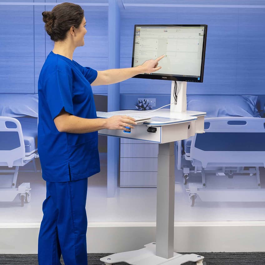 A woman in blue scrubs uses a wheeled mobile workstation, pointing at a scheduling screen beside a hospital bed in a bright clinical room.