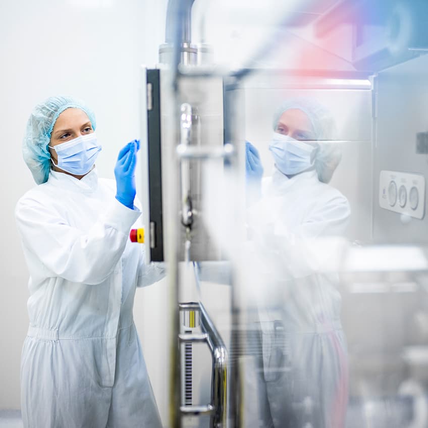 A lab technician in a white coat, blue mask, gloves, and hairnet inspects a glass panel in a bright, sterile cleanroom with control buttons visible.