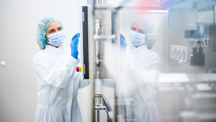 A lab technician in a white coat, blue mask, gloves, and hairnet inspects a glass panel in a bright, sterile cleanroom with control buttons visible.