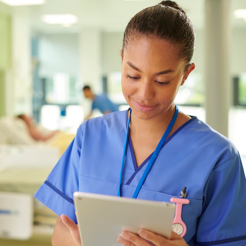 A nurse in light blue scrubs reviews a tablet beside a patient’s bed in a bright, modern hospital, with another clinician providing care in the background.