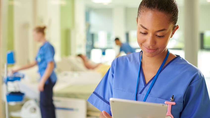 A nurse in light blue scrubs reviews a tablet beside a patient’s bed in a bright, modern hospital, with another clinician providing care in the background.