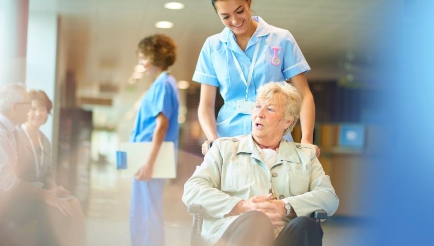 An elderly hospital patient in a wheelchair is being pushed by a young female nurse. Nurse chatting to patients in the background.