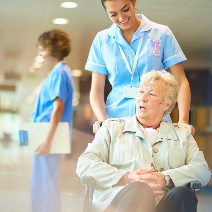 An elderly hospital patient in a wheelchair is being pushed by a young female nurse. Nurse chatting to patients in the background.