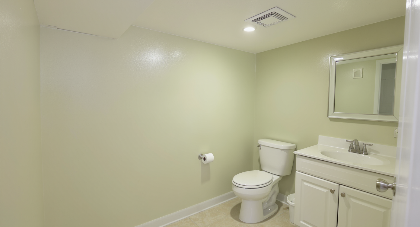 Cozy basement bathroom with pale green walls, smooth white ceiling, white toilet and vanity, GFCI outlet, exhaust fan, and light beige non-slip tile flooring.